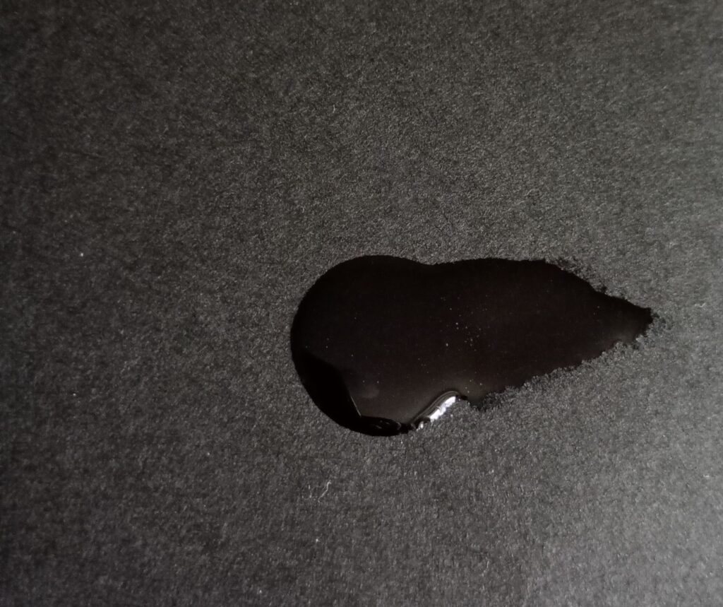 MAC リップガラスクリアを黒い紙にのせたスウォッチ。ビニールのような強いツヤと粘度が分かる質感。